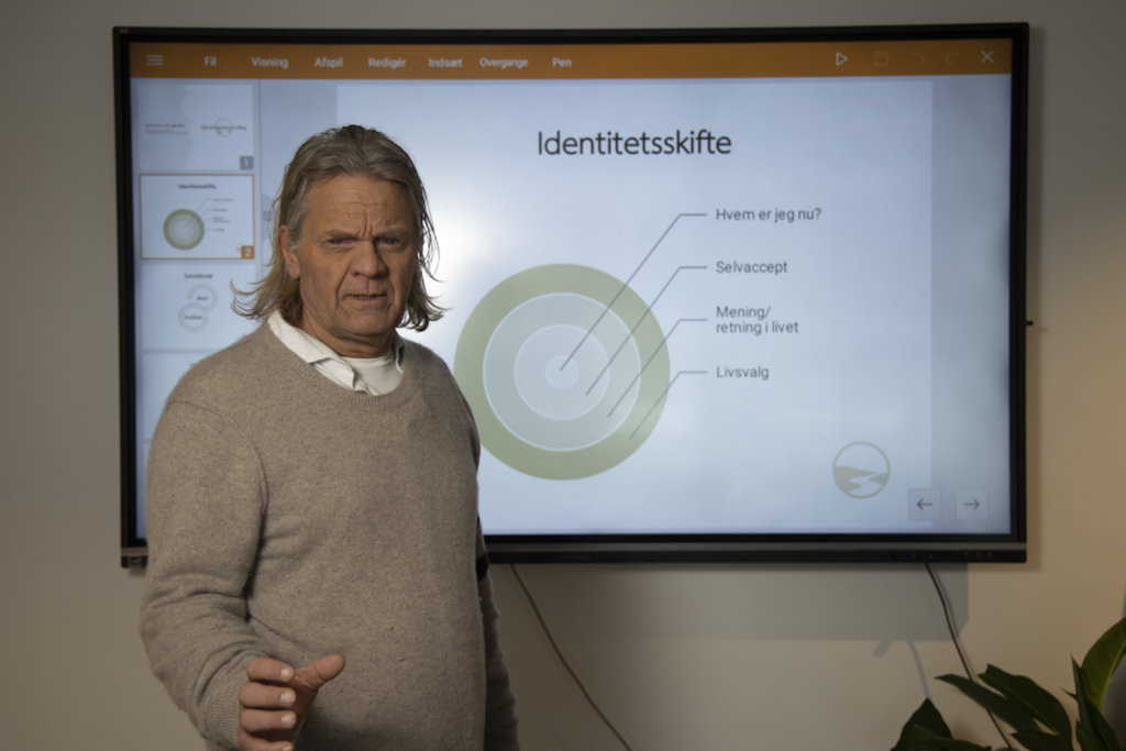 Older man in a beige sweater stands beside a large presentation screen displaying a slide titled 'Identitetsskifte' with concentric circle diagrams behind him.