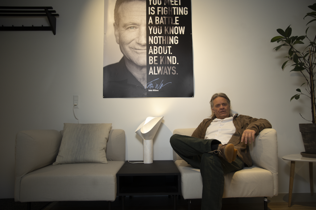 Older man relaxing on a light gray sofa in a living room, with a large black-and-white poster on the wall.