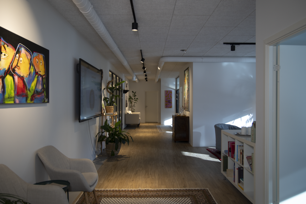 Modern office hallway with abstract wall art, plants, and a seating area along the left wall; warm wood floor and overhead track lighting.
